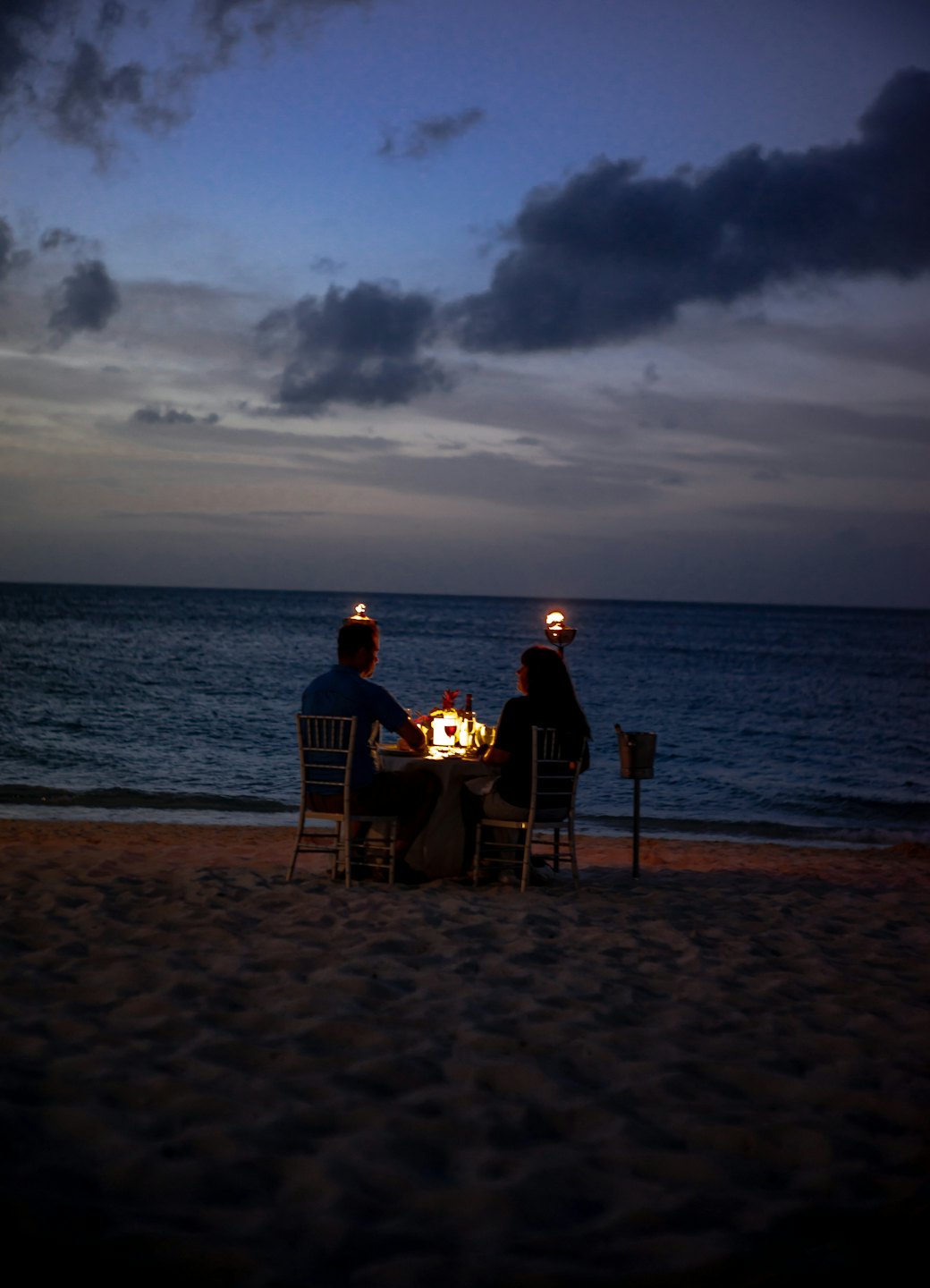 Couple enjoying a romantic candlelit dinner on a beach at dusk.